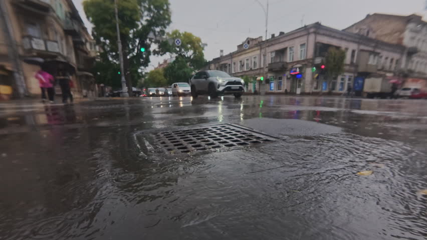 Real time, raindrops fall into autumn puddles and flows in drainage system, with cars driving across an urban intersection, people walking along street, scene of city life during rain in background