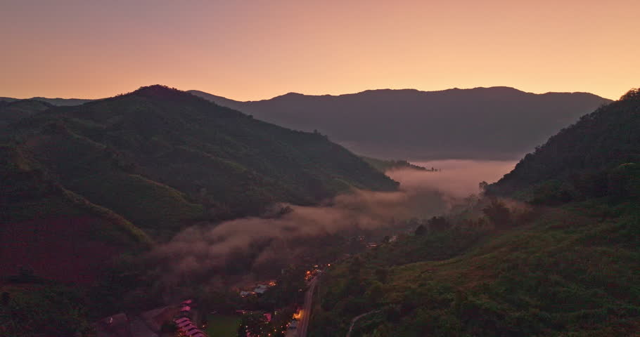 A serene aerial view of a mountain valley at sunrise, with soft mist drifting through the hills and warm lights glowing from a small village below. The scene captures the peaceful atmosphere
