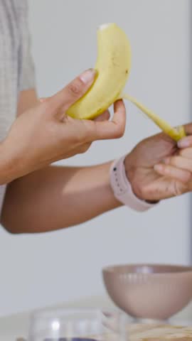 Vertical video: Peeling woman gray top gripping banana pulling peel for bowl in kitchen, copy space. Hand, fruit, closeup, vertical, countertop, minimalist, vertical video