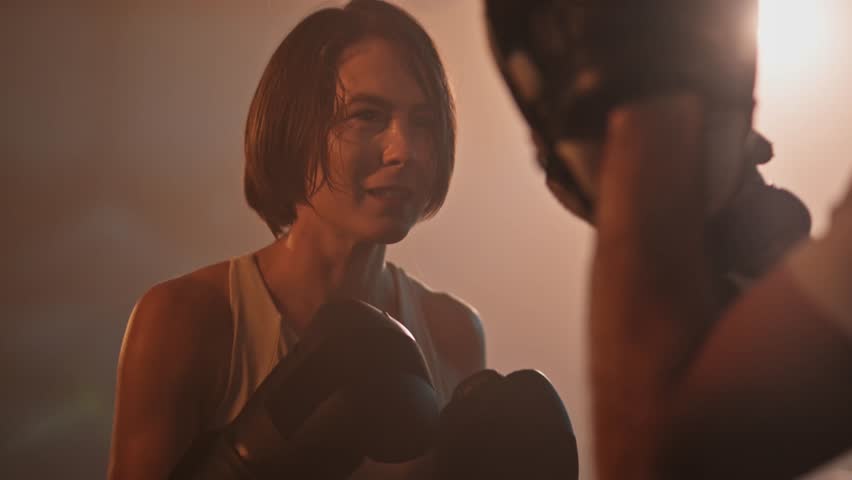 Confident woman with short hair practicing boxing punches with her sparring partner. Intense fitness training in a dark, hazy gym