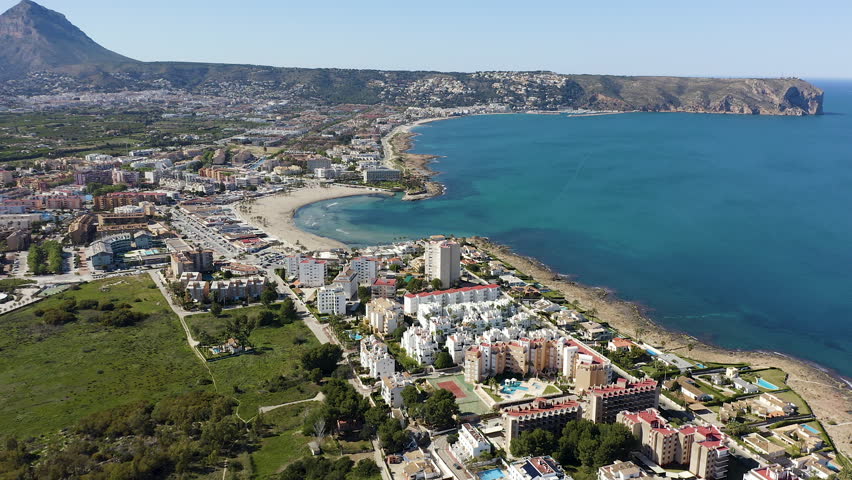 Aerial view of semicircular Arenal beach in Jávea, Alicante, Spain. Sandy shoreline with calm waters and entrance channel to marina, surrounded by urban coastal landscape. 