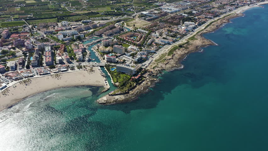 Aerial view of semicircular Arenal beach in Jávea, Alicante, Spain. Sandy shoreline with calm waters and entrance channel to marina, surrounded by urban coastal landscape. 