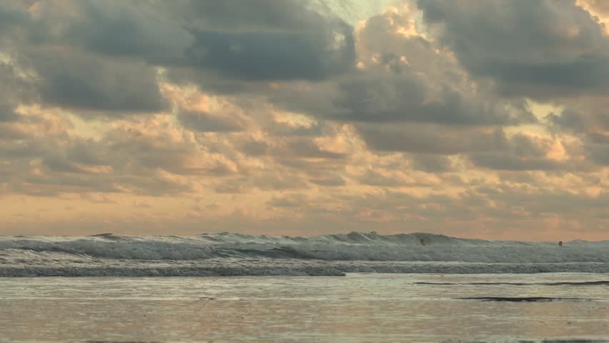 Cinematic wide shot of ocean waves crashing on a beach at sunset in Costa Rica under a dramatic cloudy sky with silhouettes of people swimming in the distance