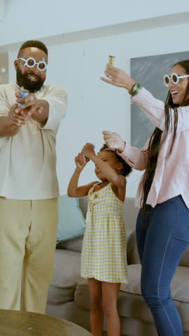 Vertical video: Diverse family popping confetti, spinning daughter, placing glasses at home for fun. Celebration, joy, playful, connection, domestic, vibrant, vertical video