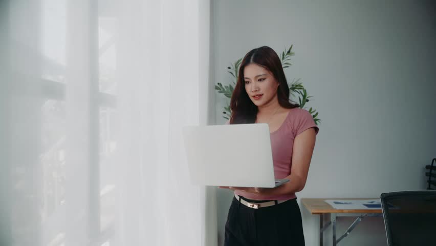 Young Asian business woman working on a laptop computer while standing near a bright window in a contemporary office setting