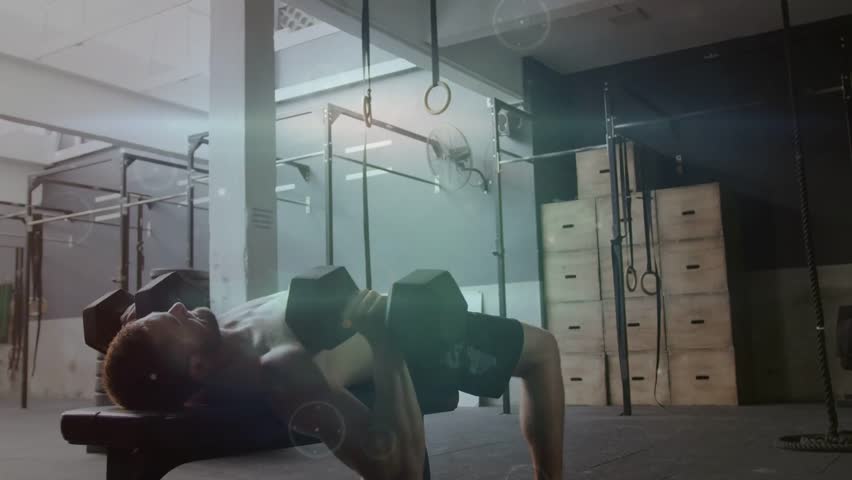 Man lying on bench pressing dumbbells from chest, building strength, light flares accenting motion. Gym, weights, lifting, training, muscular, rings, rig