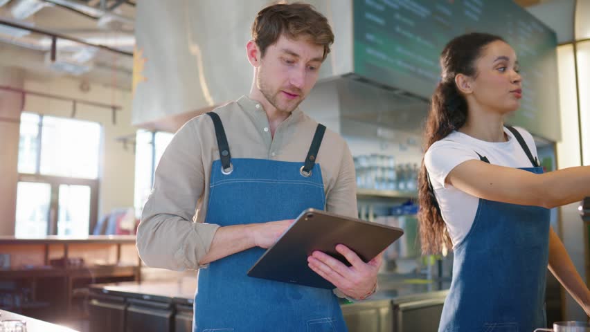 Cafe workers standing near coffee machine station. Female operating equipment and pressing controls. Male holding tablet and observing process. Coordinating drink preparation. Wearing aprons.