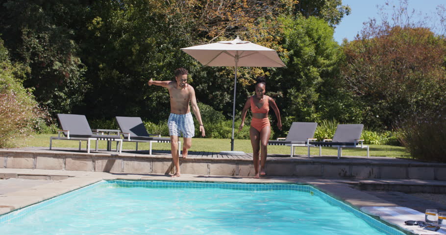 Couple stepping and running along home pool edge, launching into water for cooling, dog surfacing. Backyard, outdoor, sunny, patio, umbrella, lounge, diving
