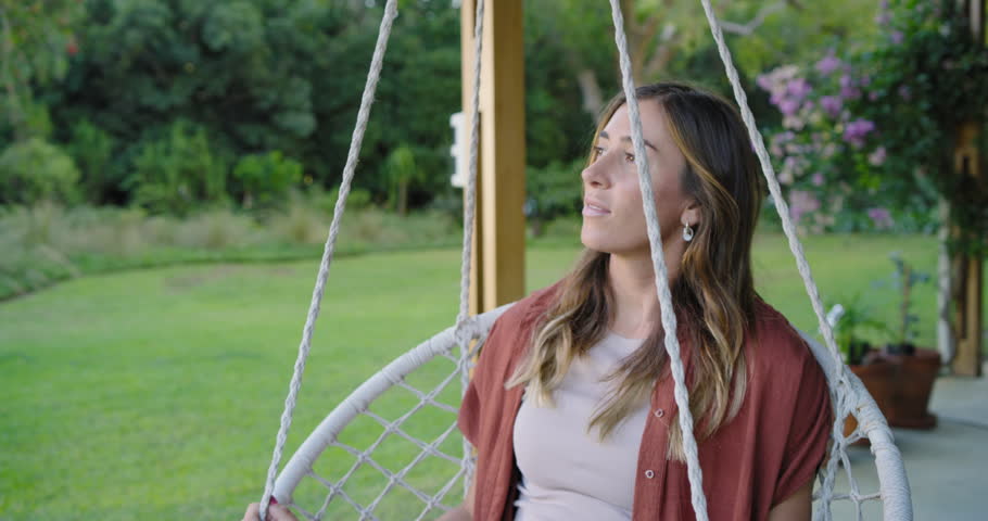 Woman wearing rust shirt sitting in macrame chair on porch holding ropes, looking up, relaxing. Outdoor, patio, garden, greenery, daylight, serene, tranquil