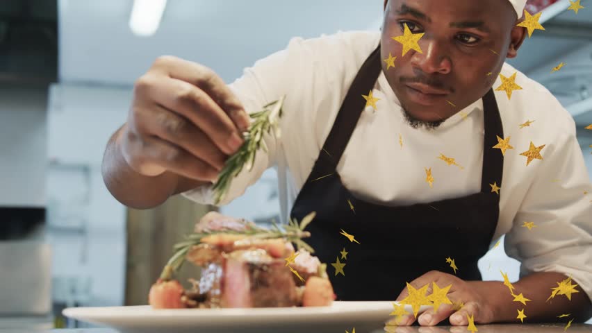 Male chef placing rosemary, lifting plated meat to pass in kitchen, confetti drifting across frame. Restaurant, plating, presentation, garnish, herb, stainless, steel