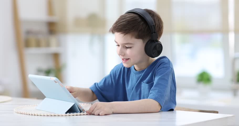 Little boy with headphones using modern tablet at white marble table indoors