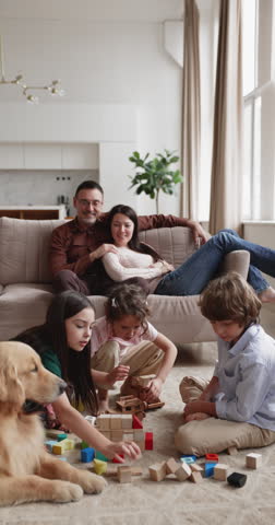 Vertical shot of large Caucasian family with golden retriever dog spending leisure time in living room, children together playing, building with wooden blocks while parents relaxing on couch at home