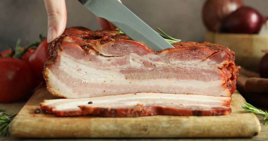 Woman cutting smoked bacon at wooden table against grey background, closeup