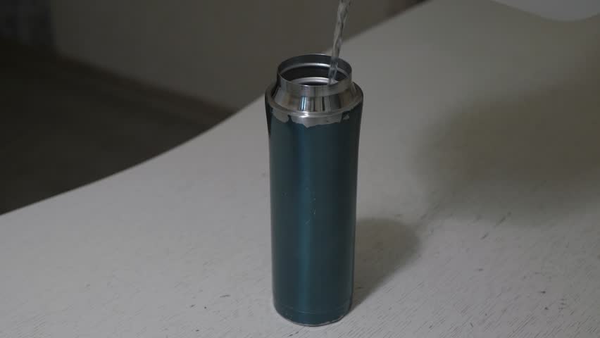 Close depiction of water flowing into metal thermos. Water filling teal bottle in dim room. Calming scene showcasing water being poured into teal metal bottle on bright surface