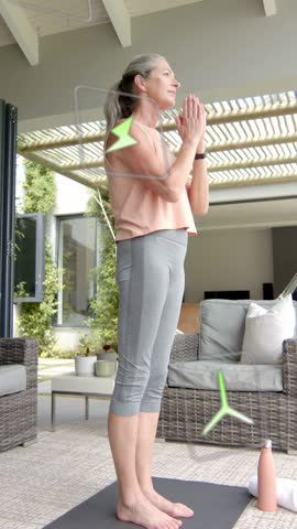 Vertical video: Woman doing flow on mat, tablet cueing energy icons drifting across her for insight. Yoga, outdoor, patio, balance, pose, wellness, vertical video