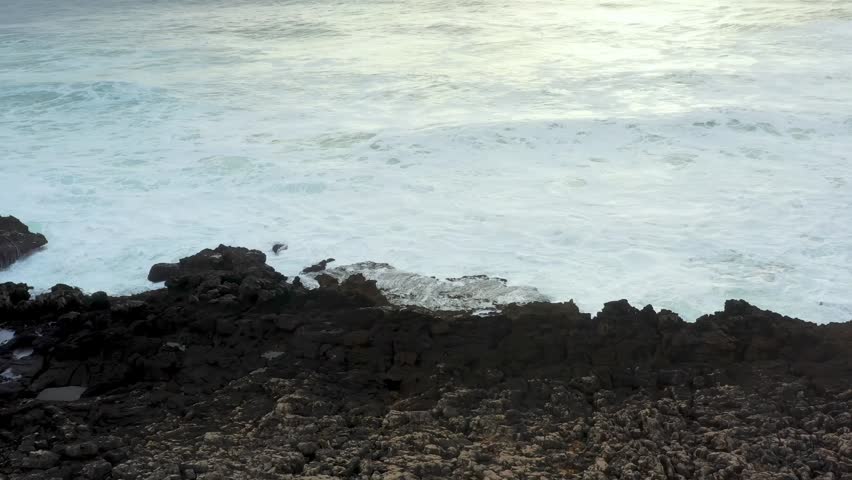 Powerful waves crash against rugged rocks as the sun sets, painting the sky in warm hues. Nature