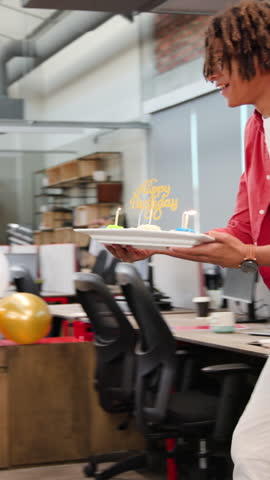 Vertical video: Diverse coworkers embracing while man placing lit birthday cake at office desk. Celebration, camaraderie, team spirit, cheerful, festive, collaboration, vertical video