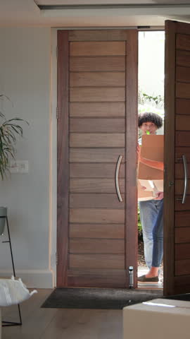 Vertical video: Diverse couple opening door and carrying boxes with plant into foyer while dancing. Romantic, celebration, home, movement, excitement, affection, vertical video
