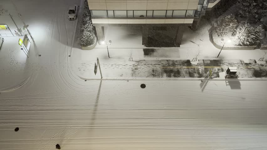 Aerial top-down night view of a snow-covered street in Sapporo, Japan during heavy snowfall. A lone person walks across the wide, freshly snow-blanketed road, casting a long dramatic shadow under the glow of streetlights. Tire tracks and footprints mark the pristine white snow, while modern buildings, a lit gas station, and snow-laden trees frame the quiet urban winter scene. The atmospheric lighting and vast expanse of snow create a serene yet cinematic winter night atmosphere.