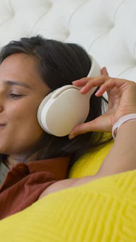 Vertical video: Raising hand, woman adjusting white headphones in bedroom to rest, wearing rust top. Cozy, headset, wireless, music, relaxed, bed, vertical video