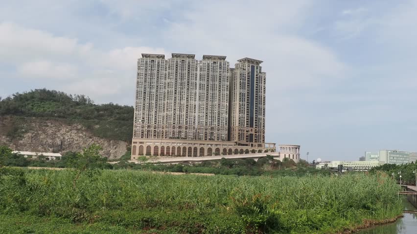 Massive residential skyscrapers tower over a lush green landscape featuring a winding canal and a wooden boardwalk pathway.