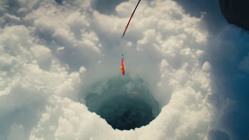 An ice fishing rod with a red lure hovers over a hole in the ice. Captures the essence of winter recreation. Ideal for outdoor activity promotions or winter sports content.