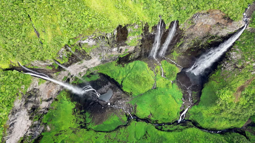 Aerial drone view of multiple waterfalls cascading down a lush green cliff into a rocky basin, surrounded by dense tropical rainforest vegetation in Madagascar