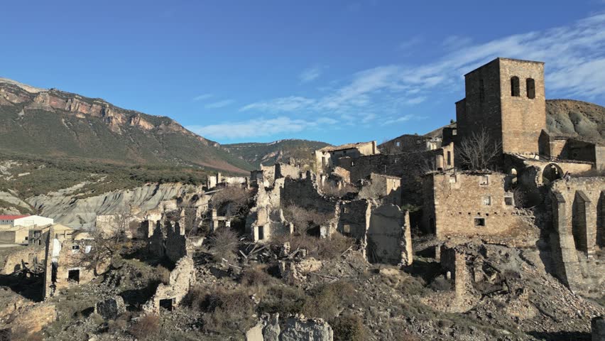 Aerial view of the medieval ruins of Esco, an abandoned ghost town on a hillside in Aragon, Spain, featuring its ancient church tower and crumbling stone houses under a clear blue sky