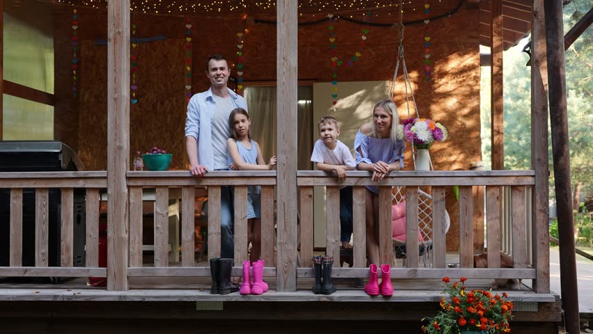 Happy Family with Two Children Standing on Wooden Porch of Country House decorated with garlands. Rubber boots lined up on deck. Rural Lifestyle, Summer Vacation Concept, Togetherness, Home Atmosphere