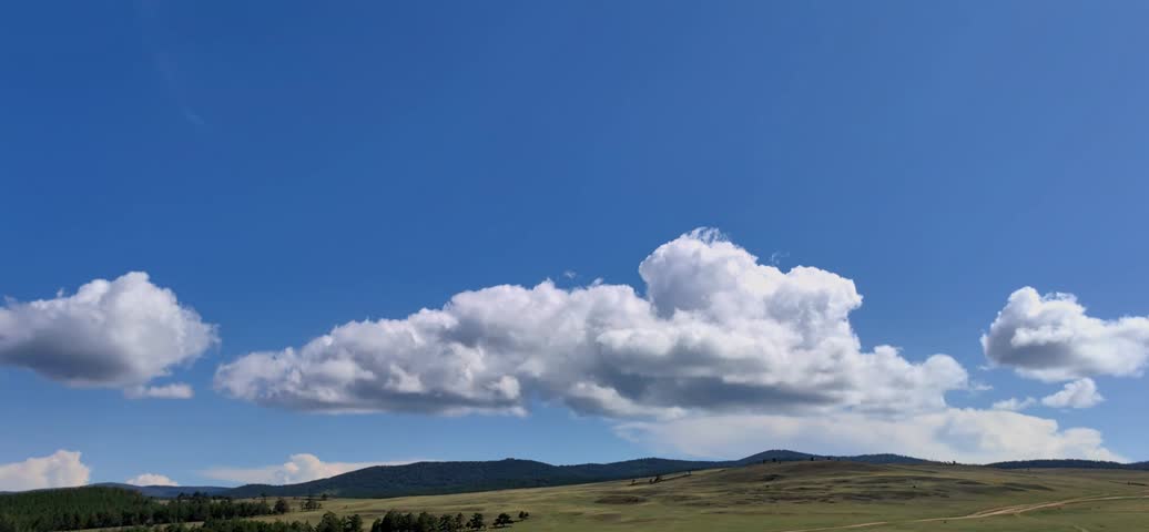 stunning timelapse fluffy white cumulus clouds moving across brilliant blue sky, with shadows sweeping over rolling green hills and distant pine forest on beautiful sunny summer day.