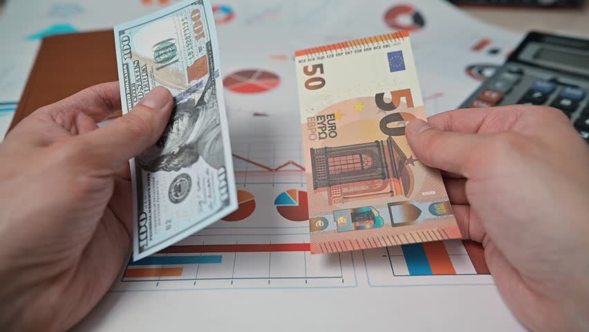 A person examines two different currency notes while seated at a desk with documents, a calculator, and graphs.