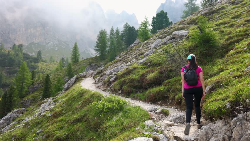 Female hiker walking along a scenic mountain path surrounded by misty peaks and alpine trees in the Dolomites, Italy. Atmospheric outdoor adventure with low clouds and dramatic mountain landscape.