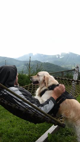 In the Carpathian Mountains, a man relaxes on a hammock with his golden retriever. They share a joyful moment surrounded by green trees and mountains in the background.