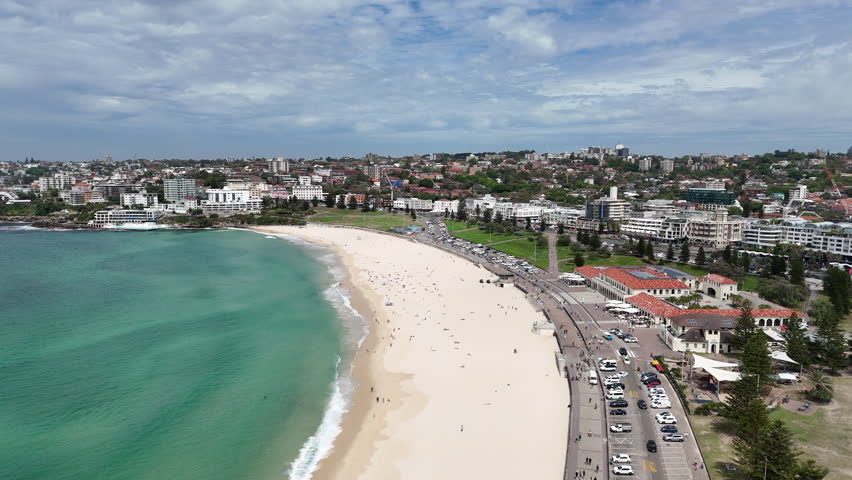 Drone footage of sandy beach coastline with turquoise water, promenade and coastal neighbourhood in Sydney.
