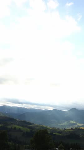 The scene shows the Carpathian Mountains in Ukraine. Lush green fields stretch across the landscape, with clouds in the sky and sunlight shining through.