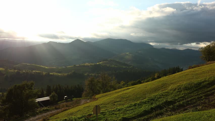 The scene shows the Carpathian Mountains in Ukraine. Lush green fields stretch across the landscape, with clouds in the sky and sunlight shining through.