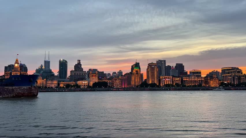 A panoramic twilight view of the historic Bund skyline in Shanghai, featuring colonial-style buildings illuminated at sunset along the Huangpu River with a large cargo ship passing by.