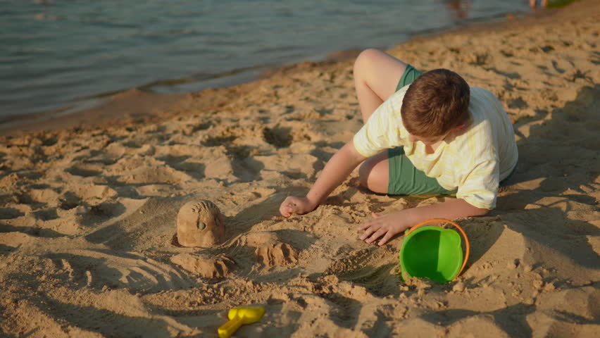 Preteen boy playing on sea shore with sand in warm sunset light. Child moving sand with hand. Development of sensory perception and creative creativity during natural interaction with nature.