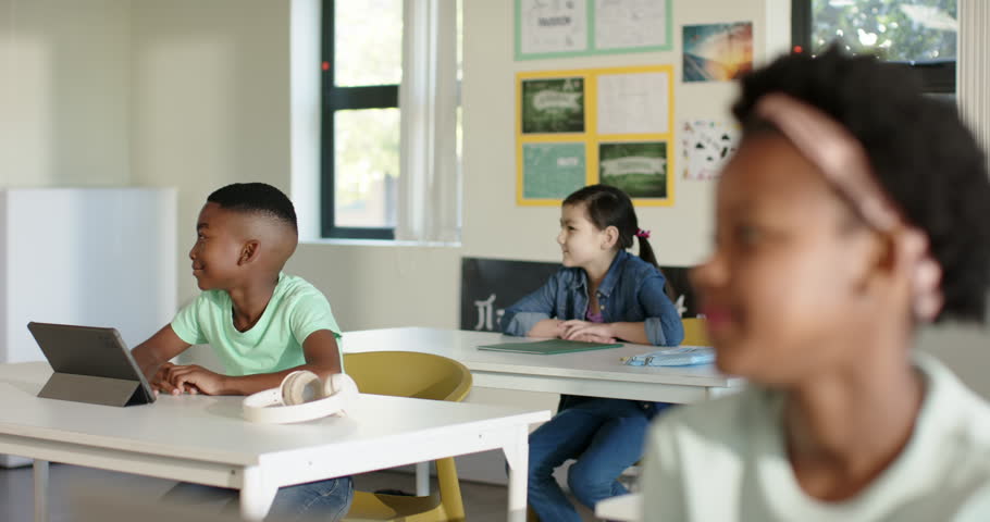 Classmates in class African American girl turning toward offscreen teacher prompt tablet headphones. Students, classroom, learning, device, window, posters, bulletin