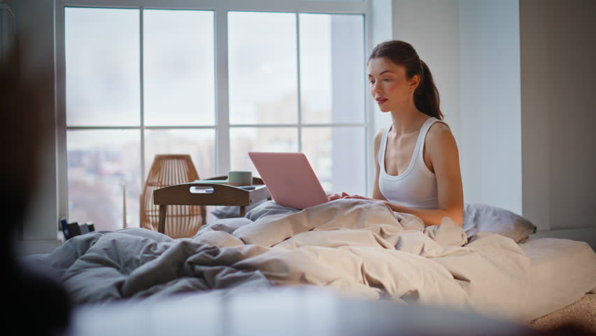 Lady closing laptop in bed finish email reading early morning. Calm beautiful woman sitting comfortable white bedding with computer on knees. Relaxed brunette freelancer working home in cozy bedroom