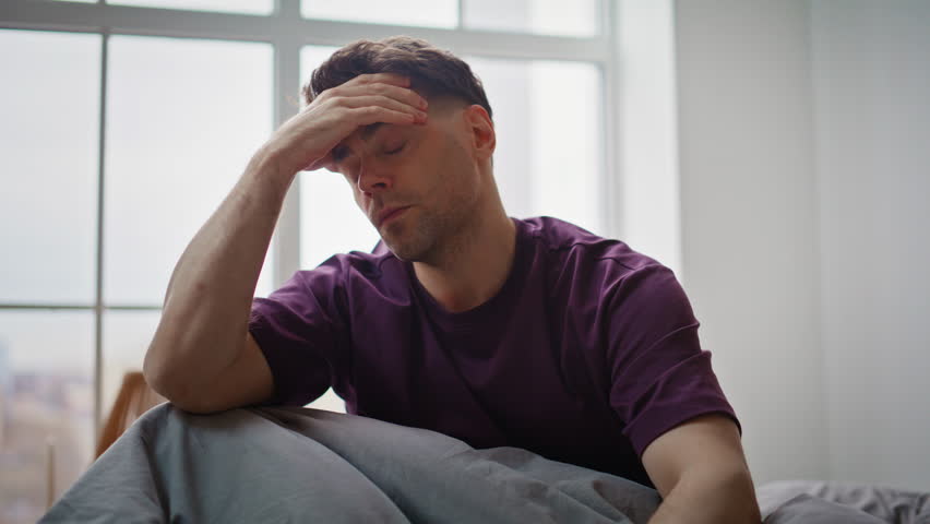 Hangover man feeling headache suffering alone at wakeup on bed closeup. Awake guy having cough in bedroom sitting on comfortable bedding. Tired brunette touching head reacting flu symptoms in bedroom
