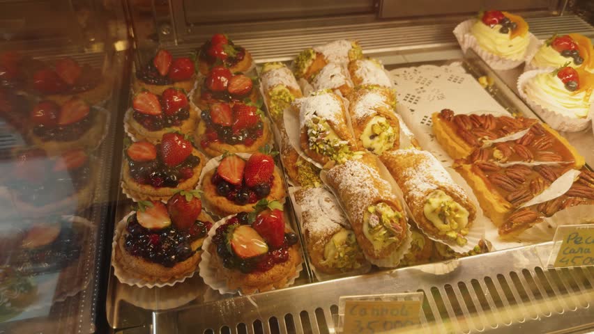 Displaying a tempting assortment of freshly baked pastries, including strawberry and berry tarts, pistachio cannoli, pecan caramel tarts, and cream meringue desserts in a bakery display case