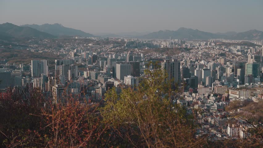 Panoramic skyline view of Seoul, South Korea, seen from Namsan Park near Seoul Tower, featuring dense urban buildings, distant mountains, and elevated cityscape perspective.