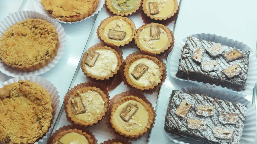 Assortment of gourmet dessert tarts, crumble cakes, and brownies artfully displayed in a bakery shop window, offering tempting individual sweet treats and baked goods selections