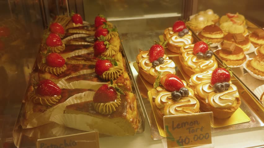 Displaying a delicious array of fresh fruit tarts including lemon meringue tarts with strawberries and blueberries, and pistachio basque slices, showcasing sweet desserts in a bakery shop window