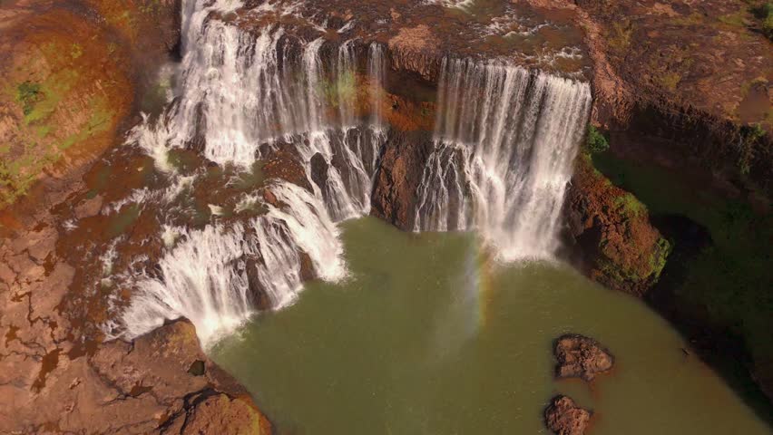Cinematic aerial drone footage of Sae Pong Lai waterfall filmed from an elevated top-down perspective, revealing the full spectacular width of the falls as multiple cascades thunder over the wide basalt ledge into the churning green plunge pool below. A faint rainbow is visible in the rising mist, created by the sunlight catching the fine spray from the powerful cascades. The rich red-orange volcanic rock surrounding the falls is fully revealed from this angle, creating a dramatic contrast with the white water and green pool. Patches of tropical forest cling to the rocky edges. This alternate angle provides unique B-roll complementing wider establishing shots of the same location. Shot in 4K UHD. Ideal for nature documentaries, adventure campaigns, and cinematic B-roll covering Laos or tropical wilderness destinations.