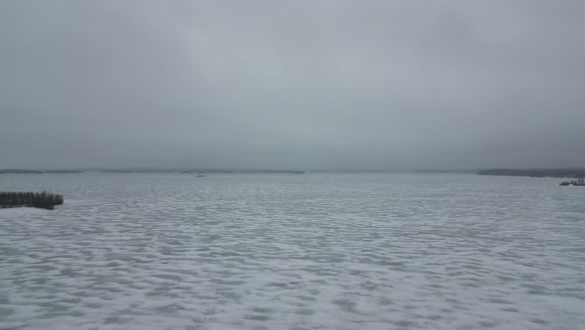 A vast aerial view of Lake Oulujärvi in Kainuu, Finland, featuring a wide frozen lake surface under heavy grey winter clouds, capturing the stark, tranquil beauty of the frozen landscape.
