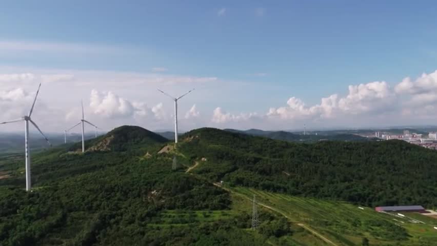 Aerial view of modern wind farm on forested mountain ridge, sustainable energy production in rural landscape, turbines rotating under bright blue sky.