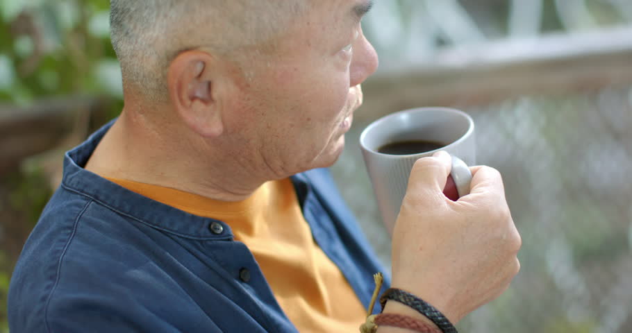 Senior Asian man sitting on porch by wood railing, sipping from gray ceramic mug, wristband visible. Outdoor, balcony, cup, beverage, calm, serene, greenery
