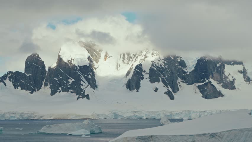 Dramatic timelapse of clouds rolling and shifting over jagged dark rocky Antarctic mountain peaks above a frozen ice shelf and dark polar ocean waters